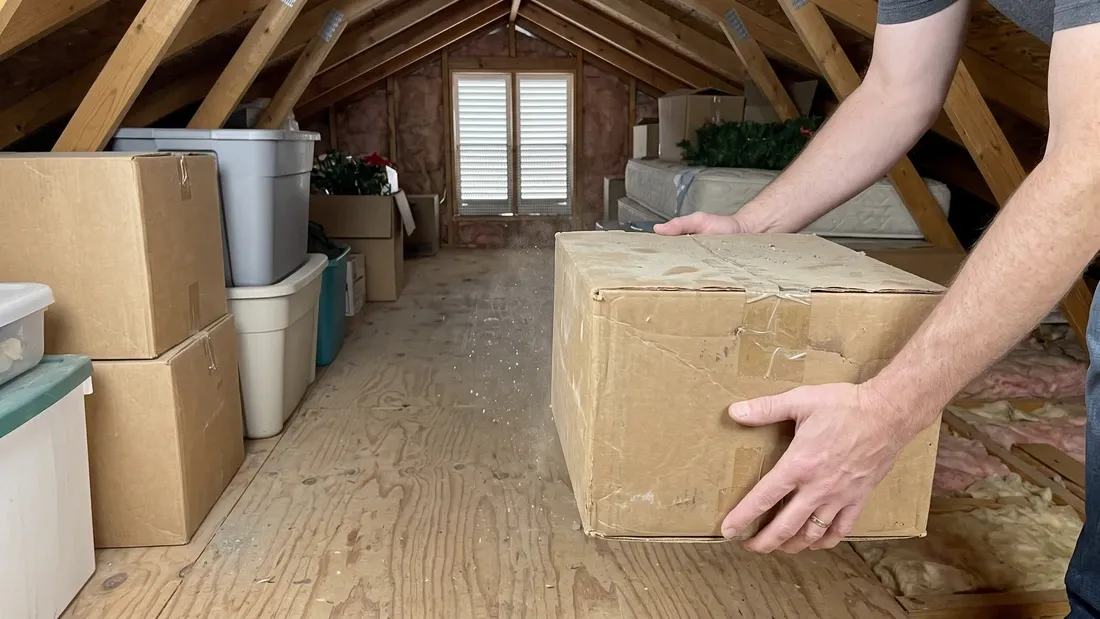 A person lifting a dusty cardboard storage box off the plywood floor of a residential attic, with more stored boxes and bins along the sides and daylight from a gable vent