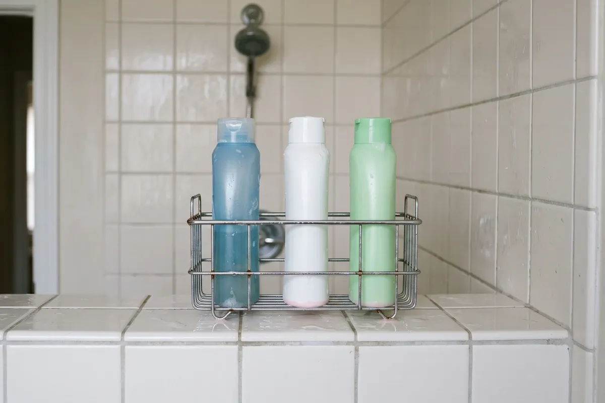 A wire shower caddy holding three plastic shampoo bottles on a tiled ledge, with a faint pink film ringing the base of each bottle where water pools.