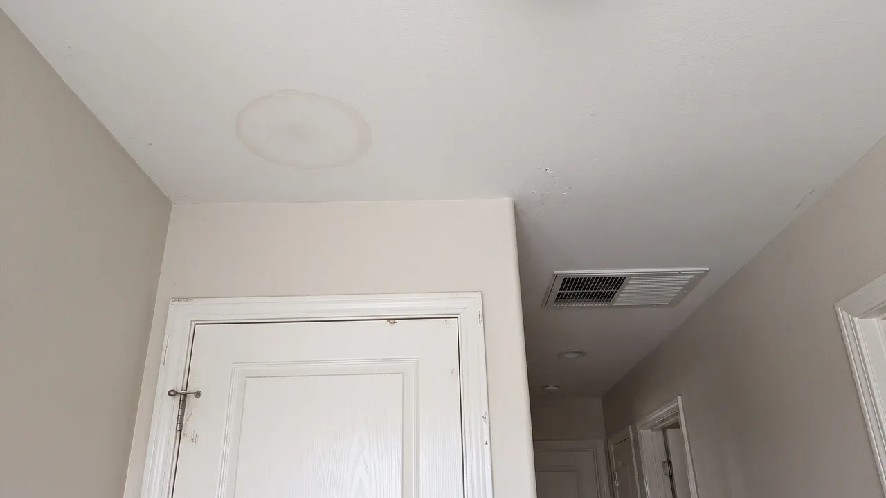 A hallway ceiling beside an air handler closet door with one faint pale tan water ring on the white paint and a supply vent nearby