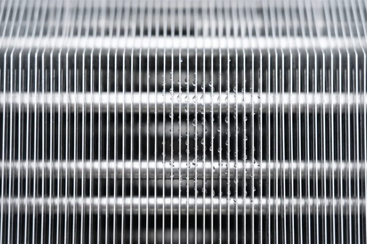 The aluminium fin face of a residential evaporator coil with scattered water droplets, the wet surface a coil lamp is aimed at