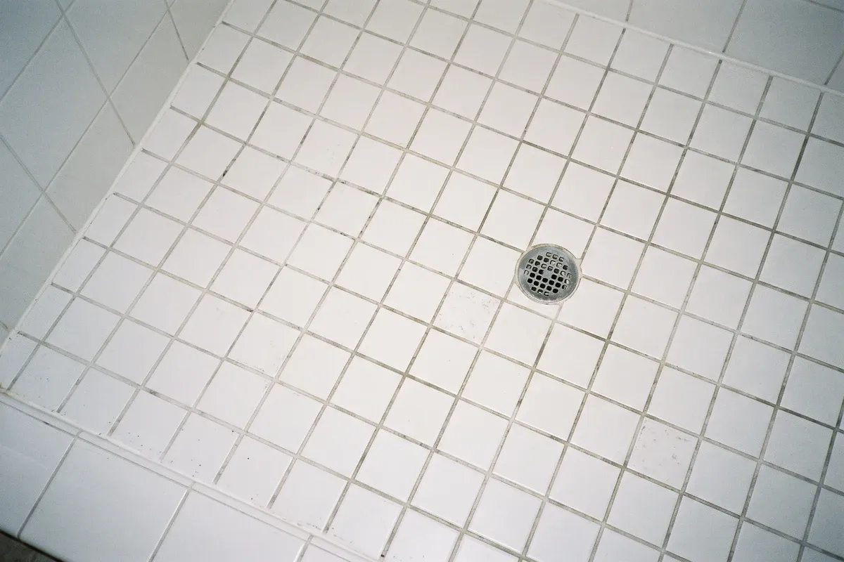 A dry white-tile shower floor with faint dark speckling confined to the grout lines between the tiles, around a stainless floor drain, with no water pooled