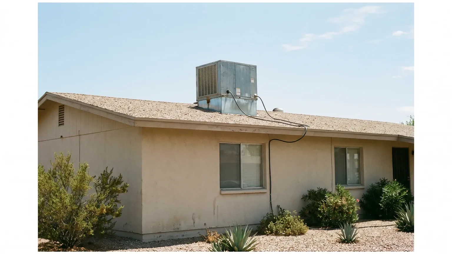 A galvanized sheet-metal evaporative swamp cooler on the flat roof of a single-story Phoenix stucco home in bright midday desert sun, water supply hose visible, desert mountains on the horizon.