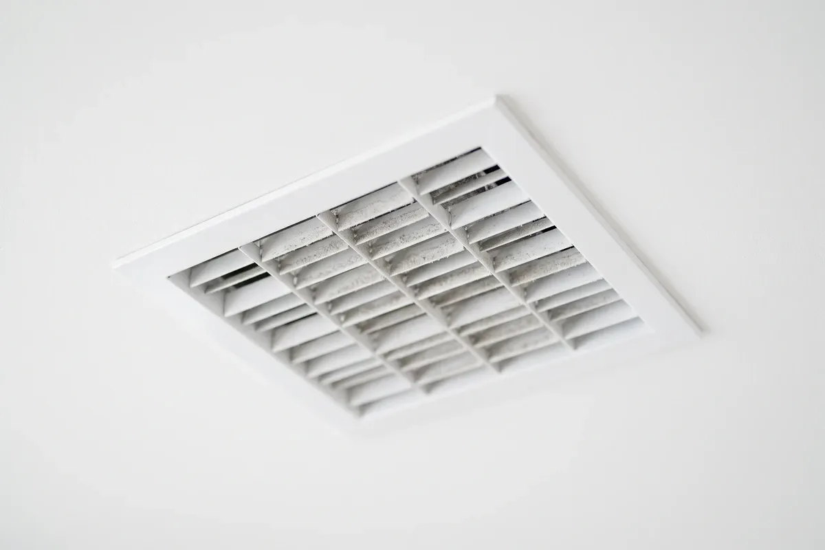 A plain white ceiling air supply register with light grey dust settled along the vanes, no gauges or displays visible