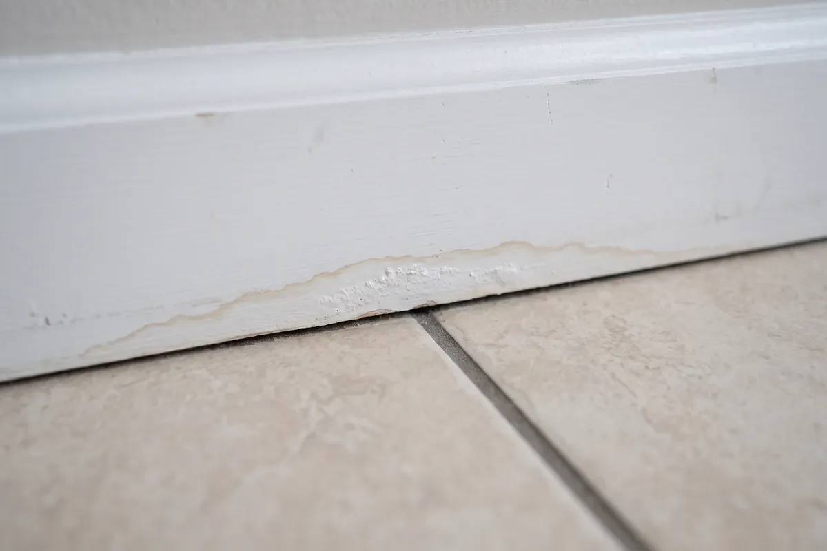 A faint light tan water stain at the base of a baseboard above a tile floor, with no visible mold growth