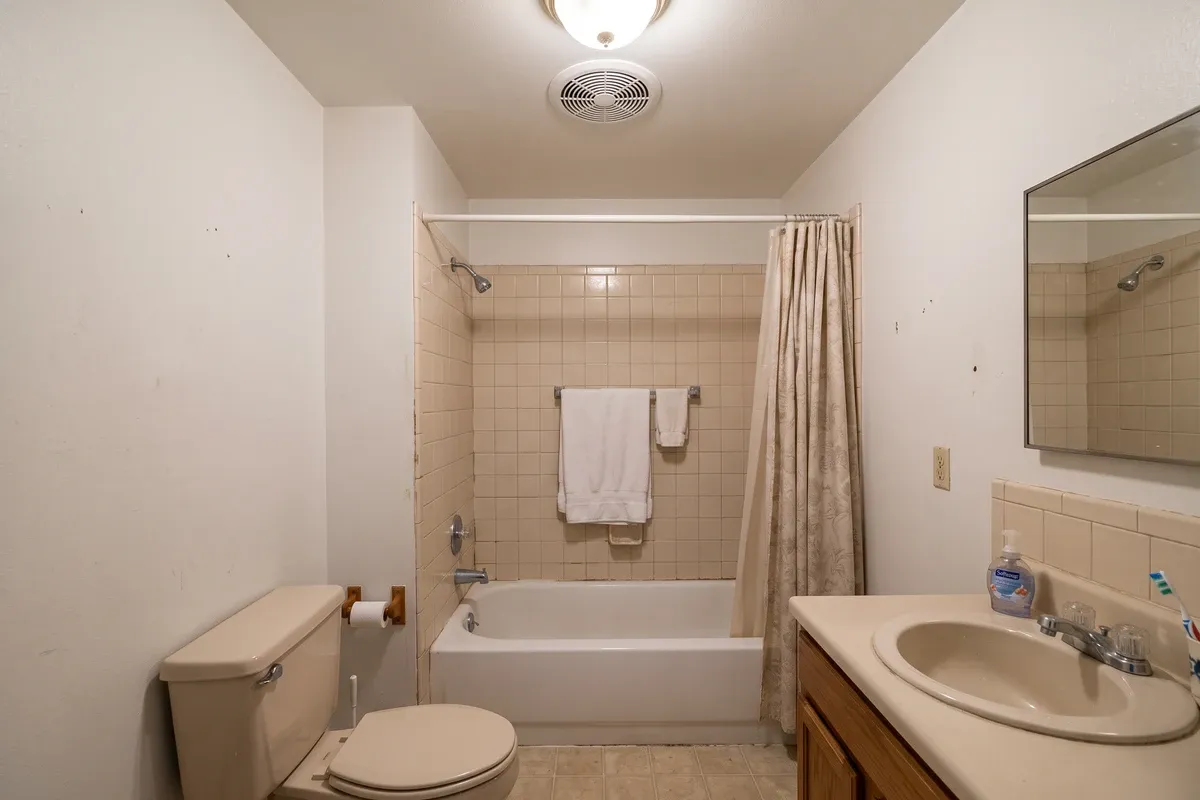 A small windowless interior bathroom with a plain white ceiling exhaust fan grille above a tub and shower curtain, beige tile and no window.