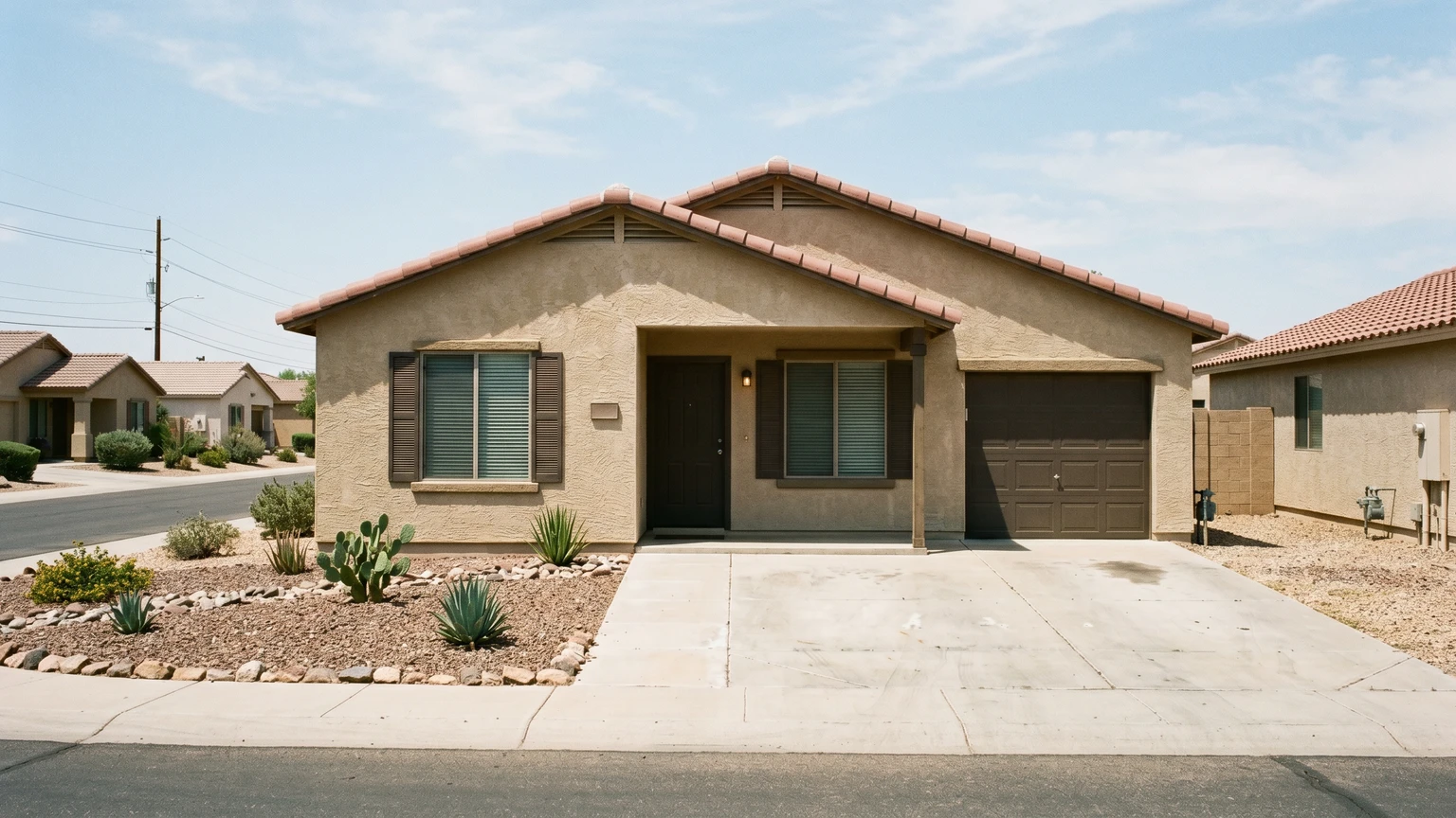 A single-story Phoenix stucco home in summer with shuttered windows, an empty driveway, and desert gravel landscaping under harsh midday sun, the home clearly unoccupied.
